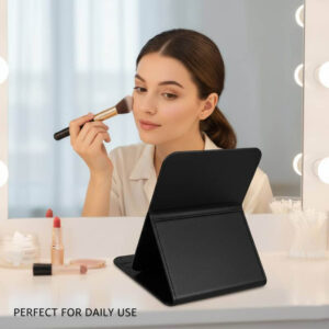 A woman applying makeup using a black portable leather makeup mirror on a vanity desk.